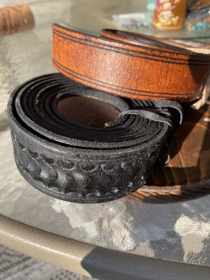 Coiled black leather belt stamped with a repeating basketweave pattern, a plain brown belt coiled behind it in the sun