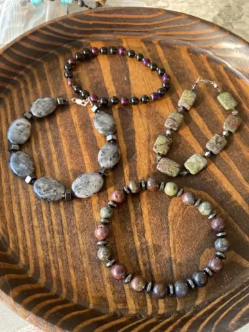 Three stone bead bracelets arranged in a wooden bowl — smoky labradorite ovals, dark rounds with purple accents, and mixed jasper rounds with metal spacers