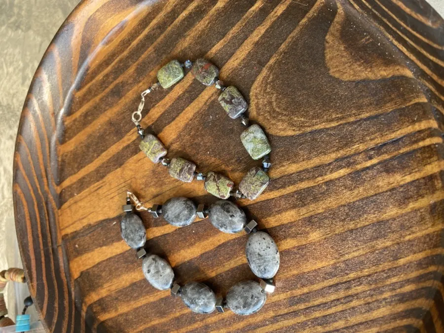 Two bracelets on dark grained wood: one of green and red dragon blood jasper rectangles, one of gray labradorite ovals with faceted metal spacers