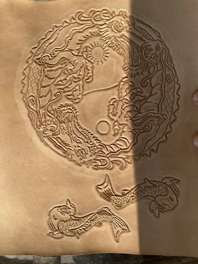 Undyed natural leather mid-carving: a circular design of a dragon and tiger curled around a yin-yang, with two koi fish carved below, before any color has been applied