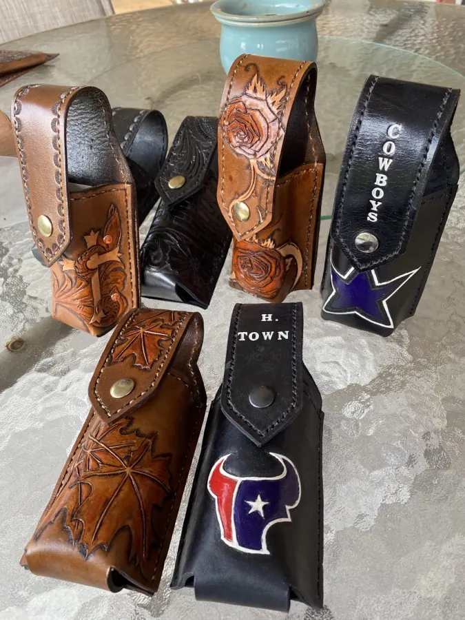 Six handmade leather snap cases: tooled cross, carved black florals, climbing roses, maple leaves, a black case painted with a blue star reading Cowboys, and one painted with a red white and blue bull reading H-Town