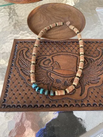 Necklace of warm sand-colored heishi beads with a cluster of chunky turquoise at the bottom, resting on the tooled leather panel