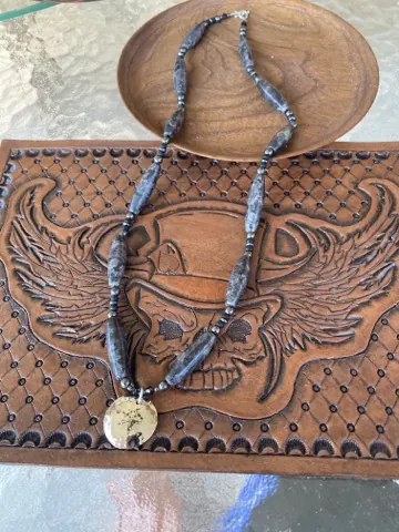 Necklace of long gray marbled stone beads ending in a hammered metal disc pendant, laid across the tooled skull panel
