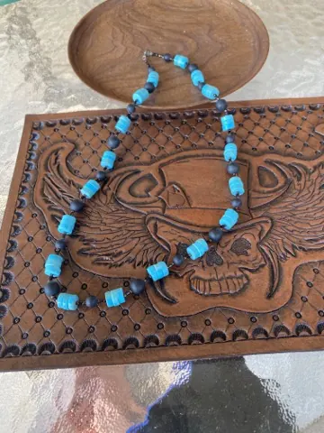 Necklace of bright turquoise cylinder beads and matte black rounds draped over a hand-tooled leather panel of a winged skull in a top hat