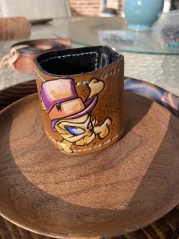 Small leather sleeve hand-painted with a cartoon character in a purple hat, stitched in teal thread, standing on a wooden plate