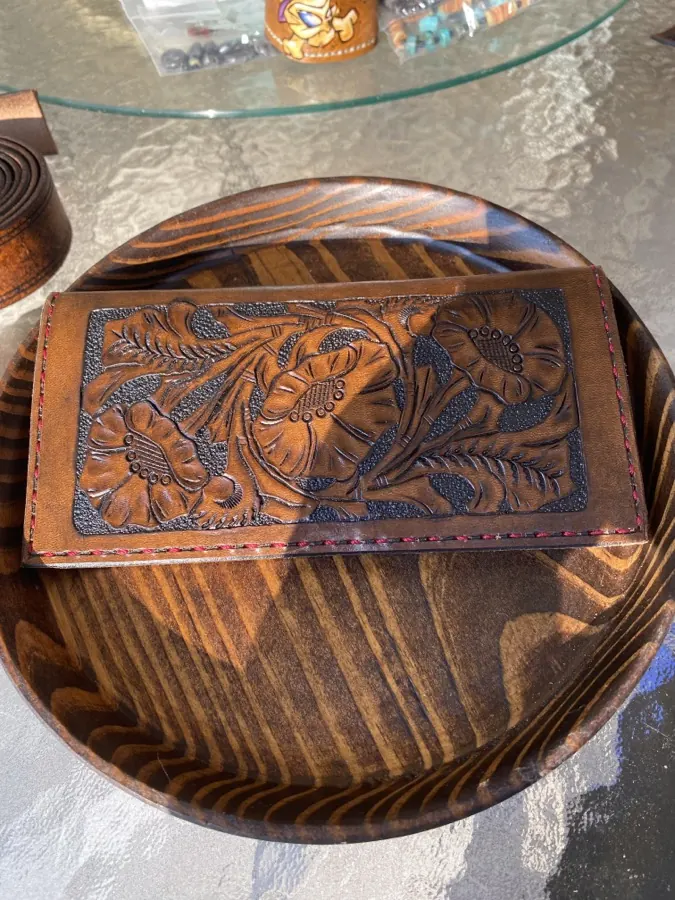 Dark antiqued leather wallet cover fully carved with wild roses and foliage, edged with red stitching, resting on a striped wooden bowl in warm sunlight