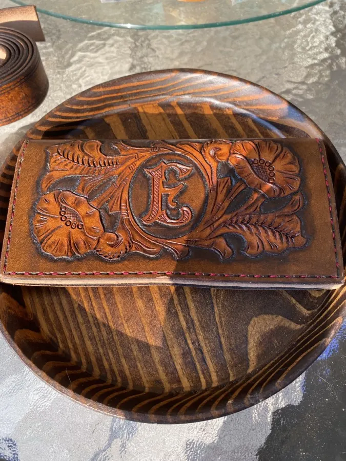 Outside of a floral-carved long wallet with a tooled letter E monogram at its center, red stitched border, warm cognac leather